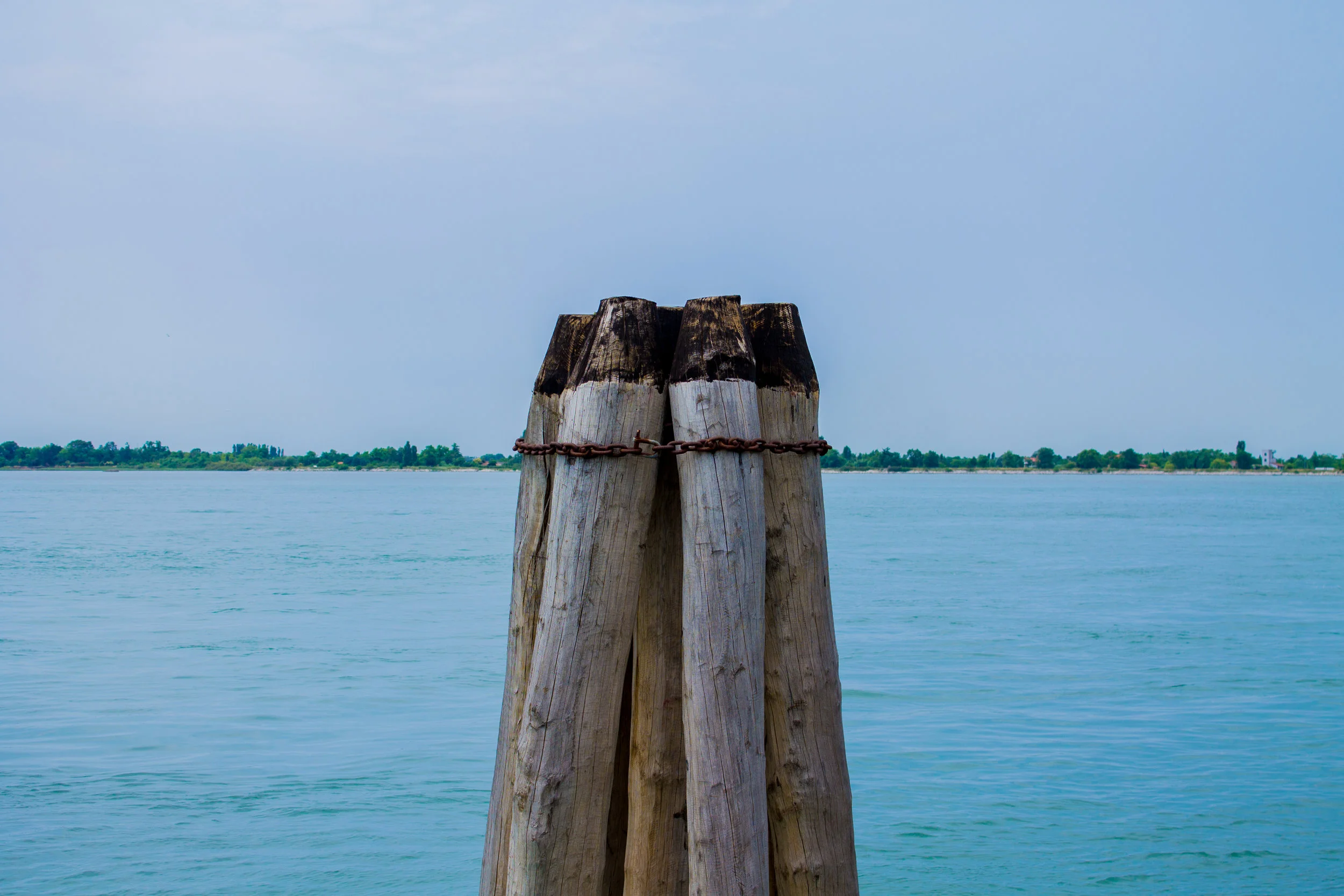 Four weathered wooden posts with a chain in front of a body of water and distant shoreline under a partly cloudy sky.