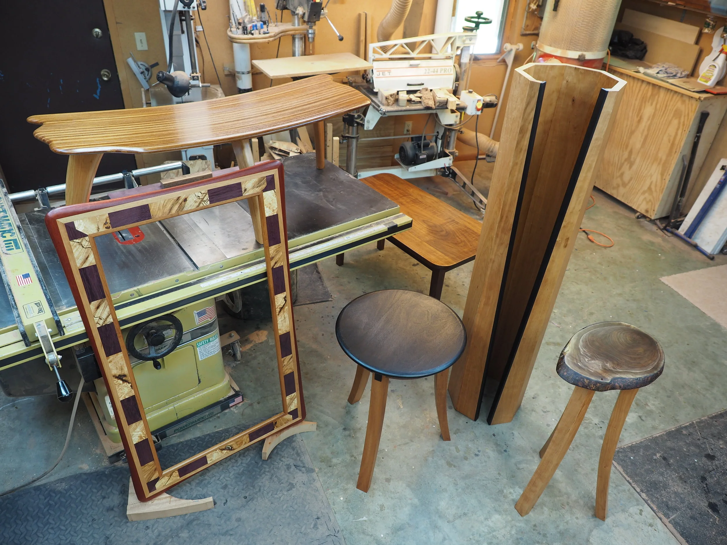 L to R: A Bayfield Collection Burl Mirror, Bent Lamination Coffee table (on the saw), 3-leg Showcase Table, Open Portal Lamp, and a small Showcase Table (live edge top).