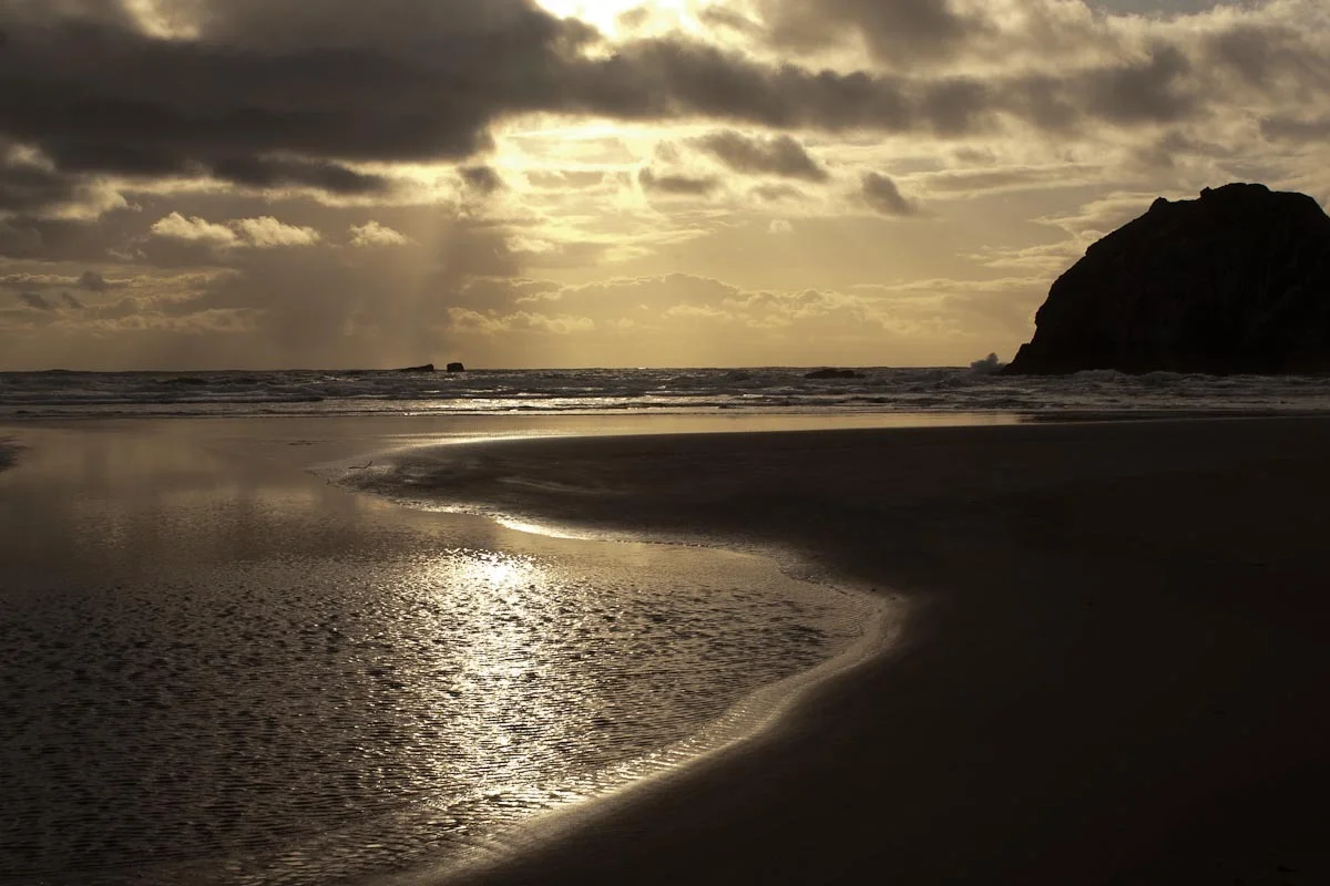 A sepia toned image from a beach at night. The sand and water form almost a yin yang shape. The sky is cloudy with sun shining through the clouds. In the far right is a rounded hill in the distance. 