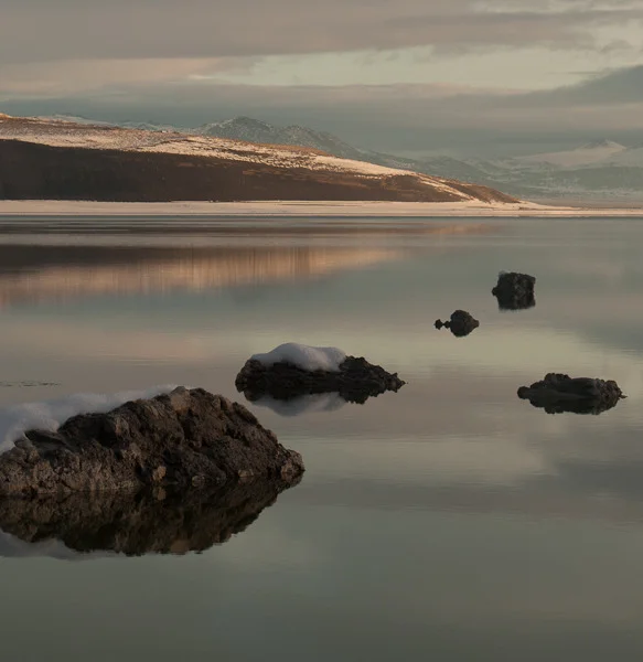  A still lake with silhouetted short tufa formations in it. There are snow covered hills in the distance. The colors are ice blue, green, and pink. The sky is cloudy.  