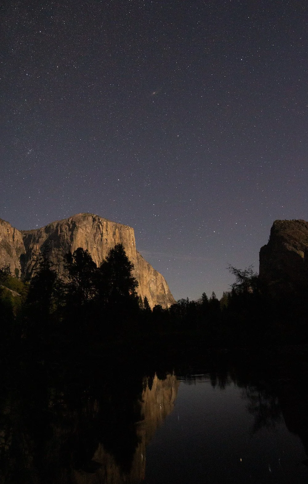  View from a lakeshore looking towards trees and a mountain. There are many stars in the sky and some lights on the mountain which may be from lights that climbers are carrying. 