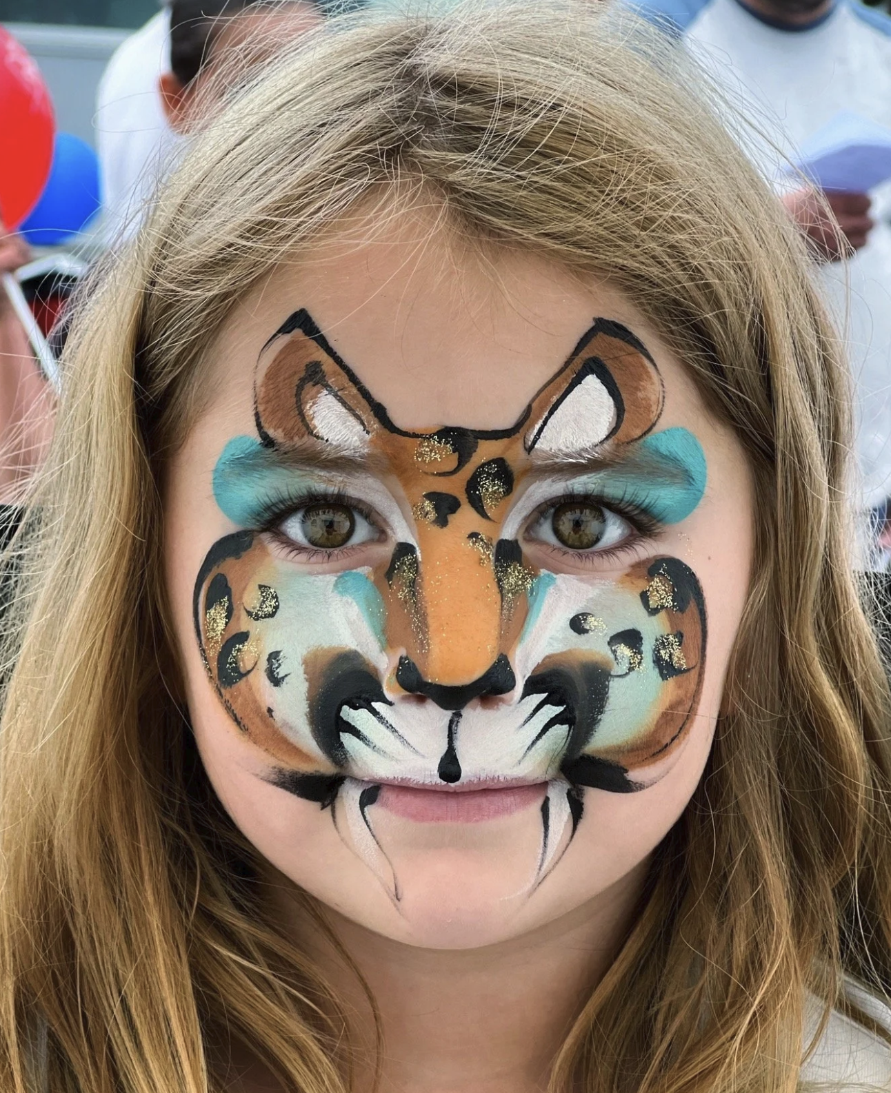 A young girl with detailed face paint resembling a tiger, including orange, white, black, turquoise, and gold accents.