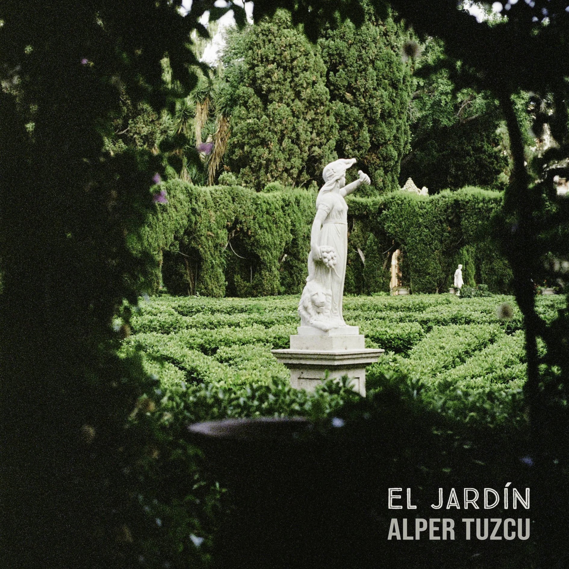 A white marble statue of a woman in a garden with trimmed bushes and tall trees, viewed through a natural frame of dark foliage. The image includes the text "El Jardín Alper Tuzcu" in the bottom right corner.