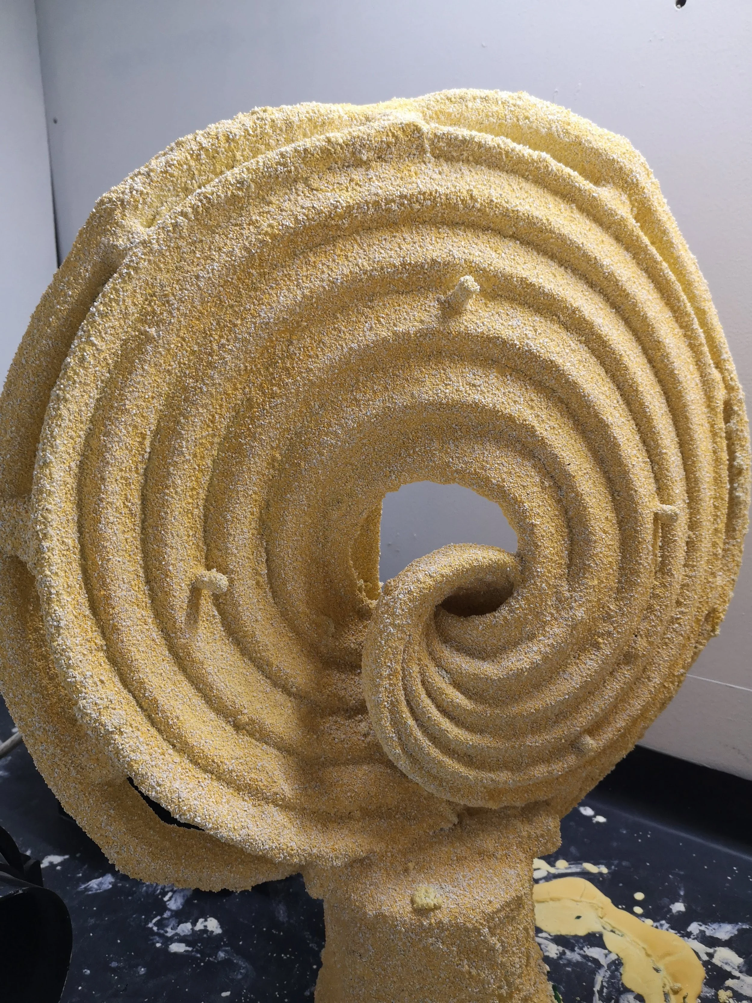 A close-up view of a textured, yellow, spiral-shaped cake with multiple layers, covered in a coarse sugar coating, with some decorative elements on top and some cake crumbs and icing on the black surface beneath.