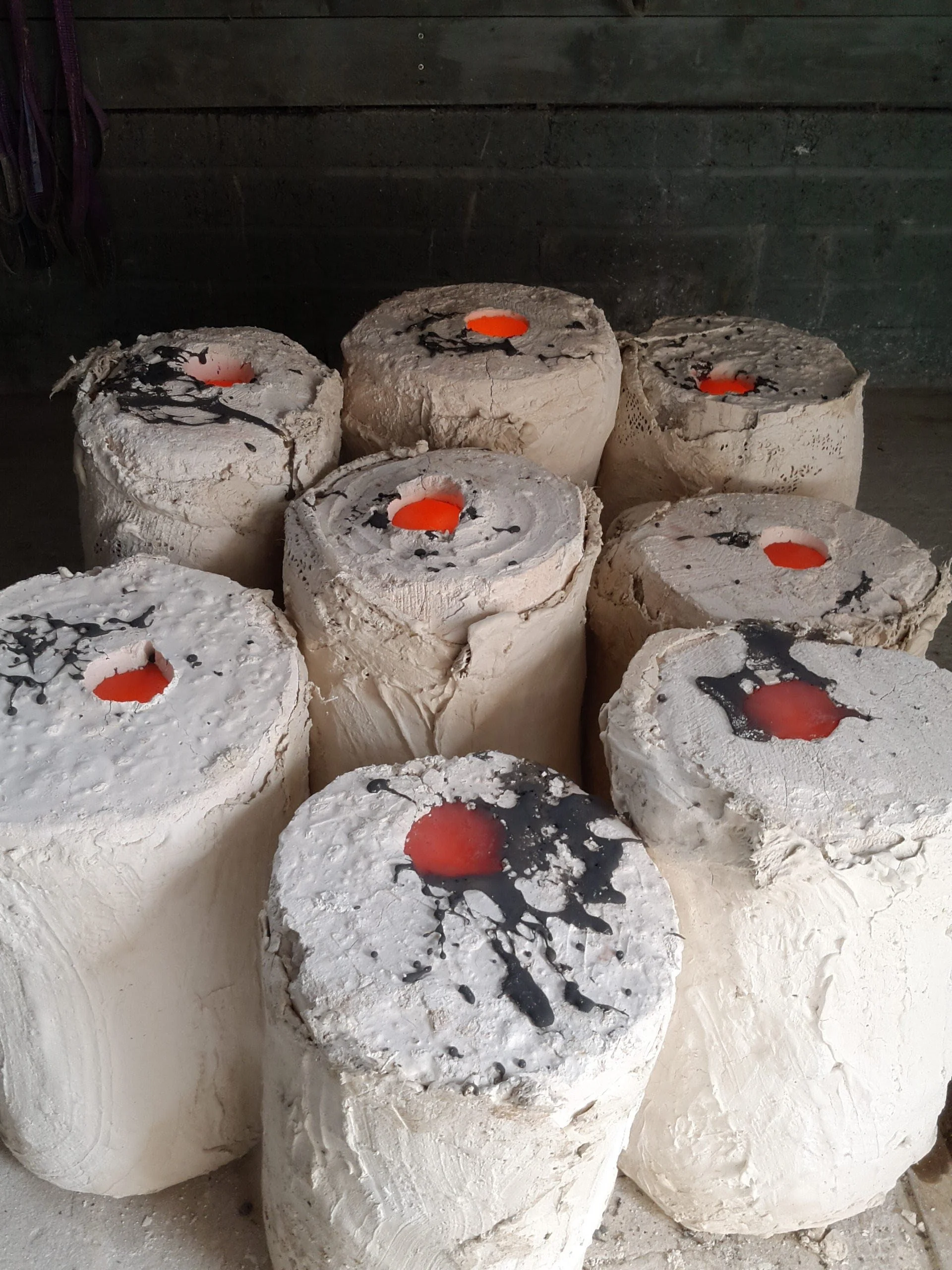 Nine large candles with white textured wax and orange centers, decorated with black splatter patterns, arranged on a concrete surface against a dark gray wall background.