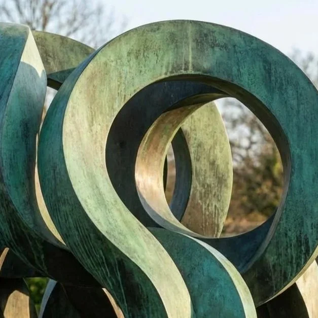 Abstract greenish bronze sculpture with looping, intertwined shapes outdoors on a stone platform, with trees in the background.
