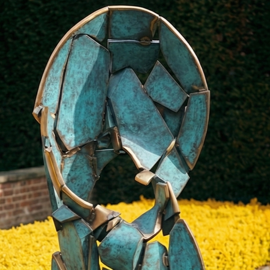 Sculpture made of blue and copper geometric shapes stands in a flower bed of yellow flowers, with a brick wall and green hedge in the background.