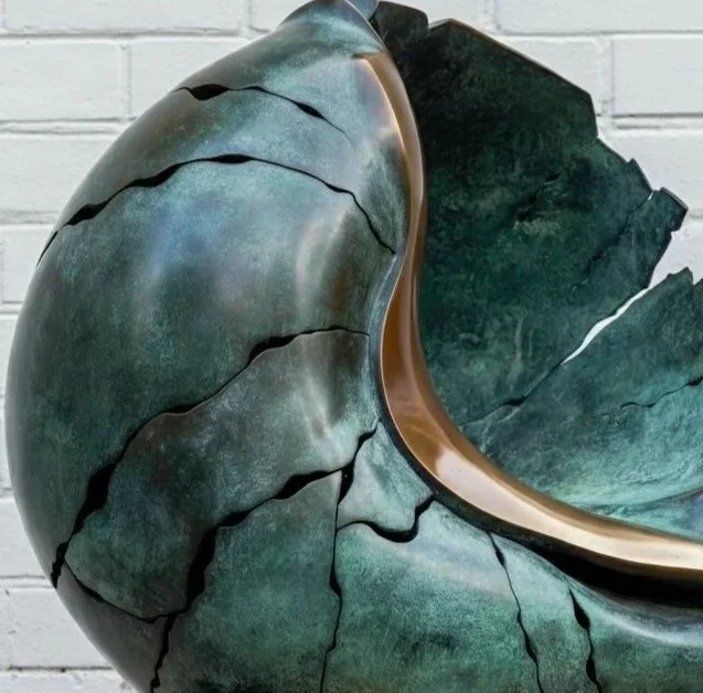 A large, abstract sculpture resembling a breaking shell or layered rock formation, made of metallic and stone-like materials, mounted on a rectangular base, set against a white brick wall.