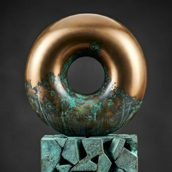 A modern sculpture featuring a large, metallic gold ring with a hole in the center, mounted on a textured greenish stone block with cracks and angular pieces, against a dark background.
