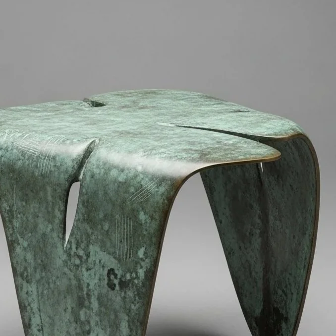 Green patina metal sculpture with curved, abstract form resembling a table or furniture piece, against a gray background.