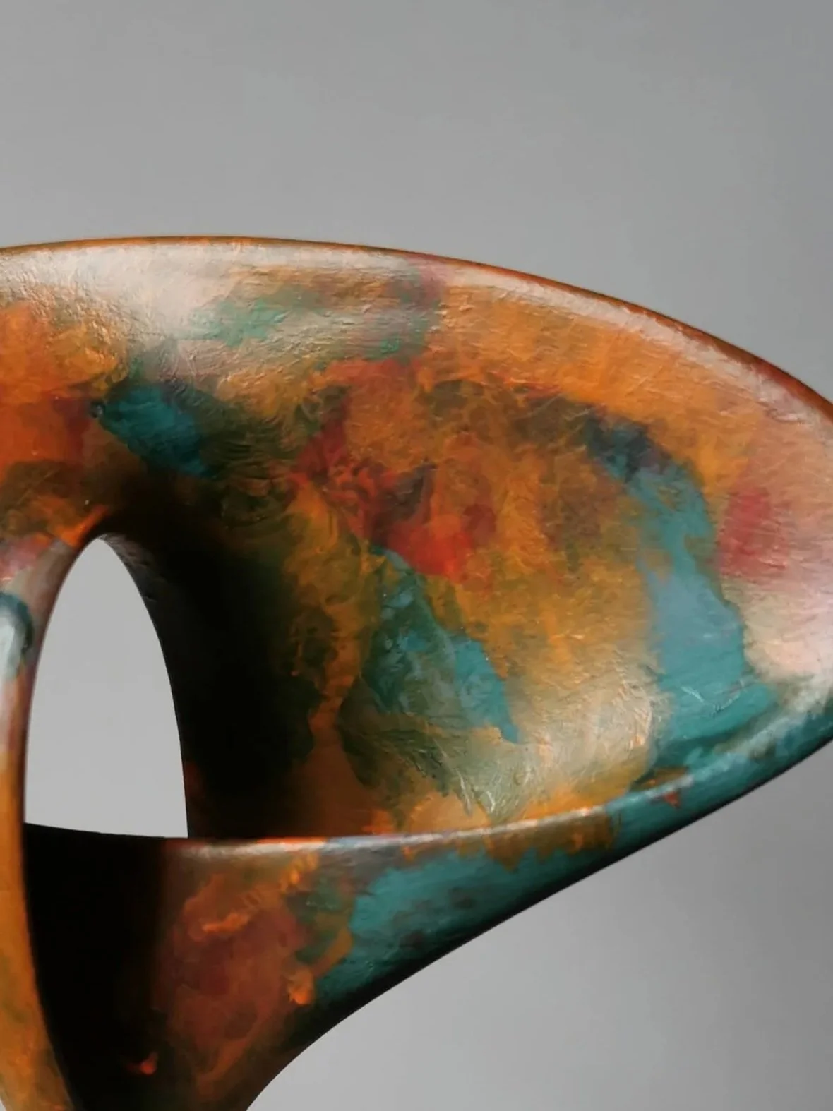 Colorful ceramic sculpture with an abstract, curved, spiral shape, placed on a white square pedestal against a plain gray background.