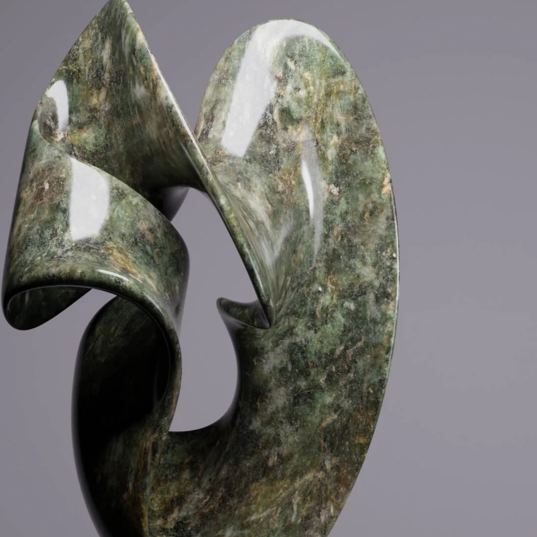A modern sculpture made of greenish marble with smooth, curved, abstract shapes, displayed on a black pedestal against a plain gray background.