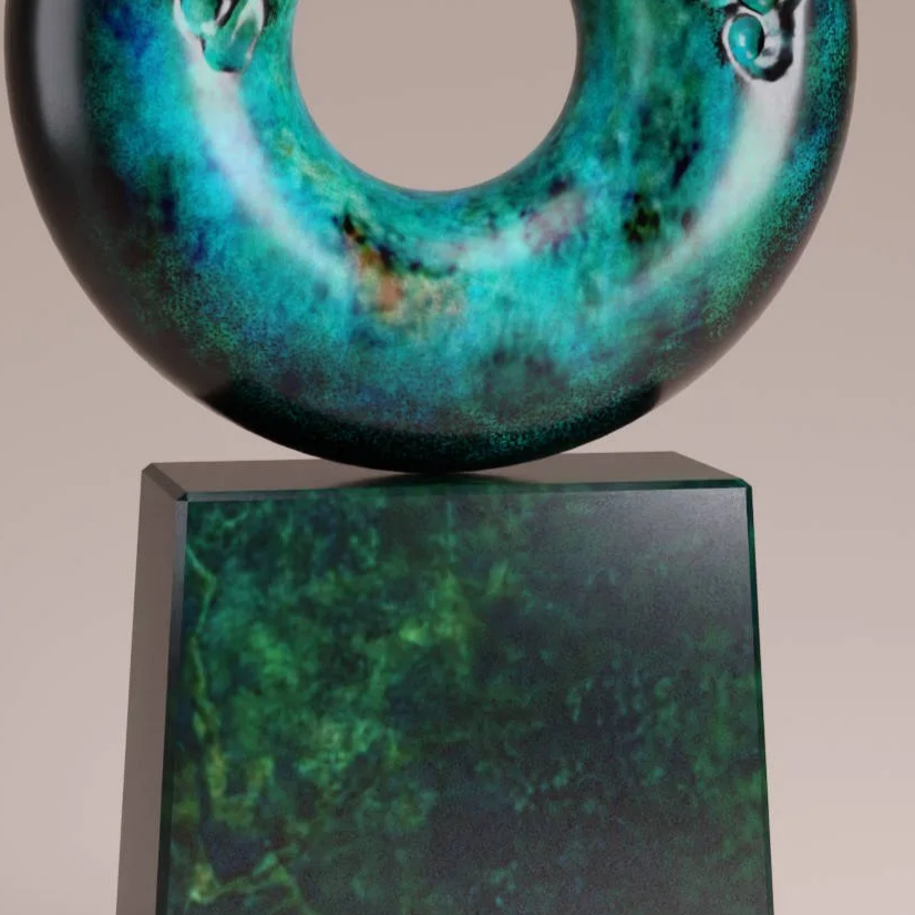 A circular abstract sculpture with a textured, greenish-blue surface, mounted on a rectangular stand, against a plain background.