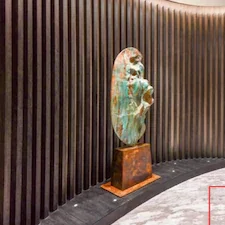 Sculpture of a woman's head mounted on a pedestal against a vertical wooden panel wall.