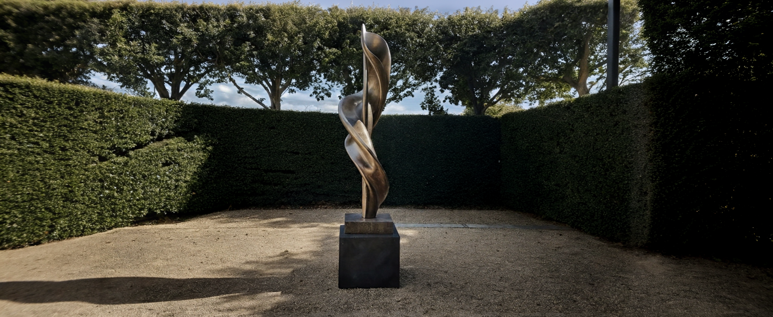 Outdoor bronze sculpture with twisting, organic shapes, placed on a square base in a fenced garden area with tall hedges and trees.