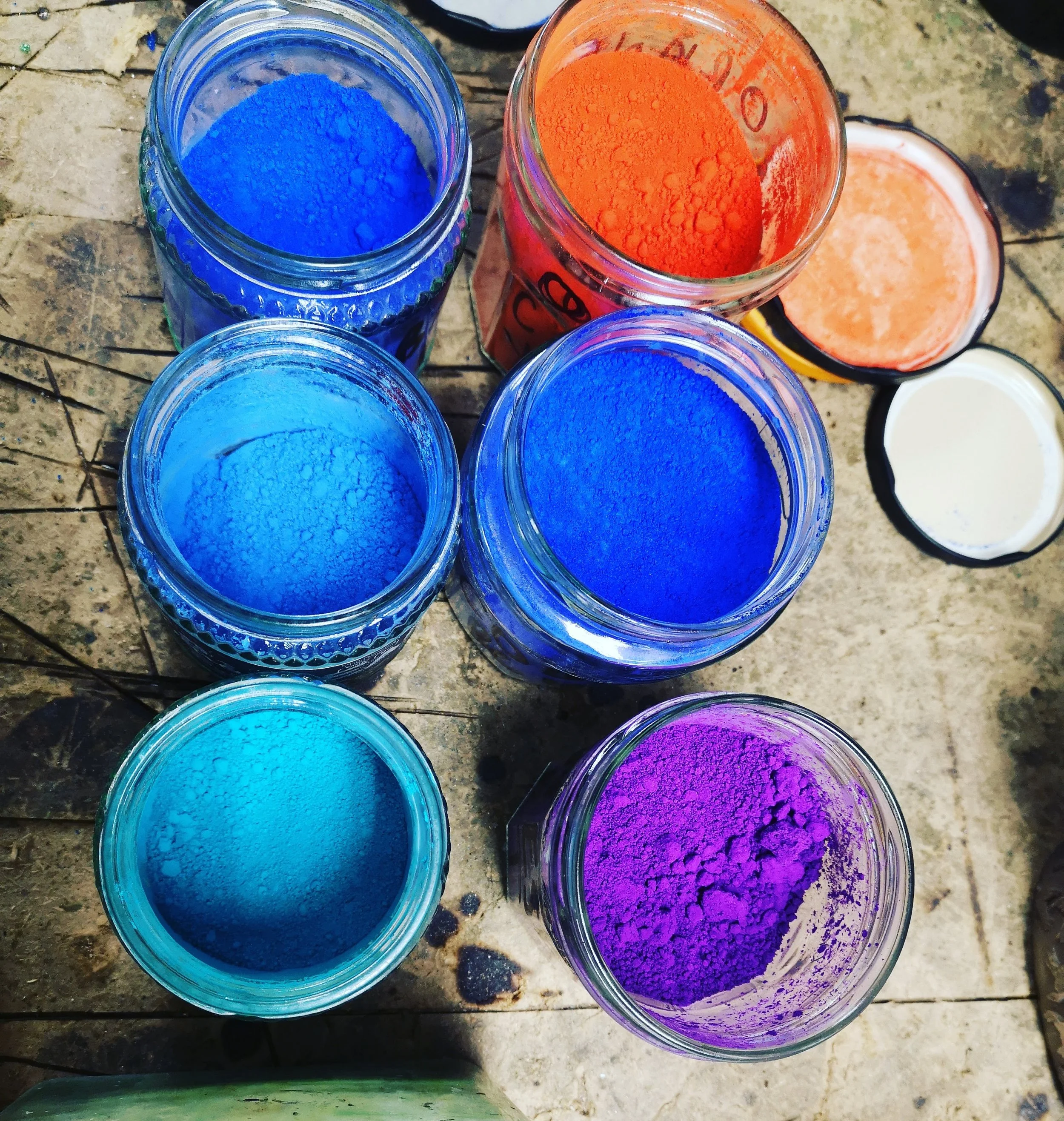 Several jars filled with brightly colored powders, including blue, orange, and purple, placed on a rough, rustic surface.