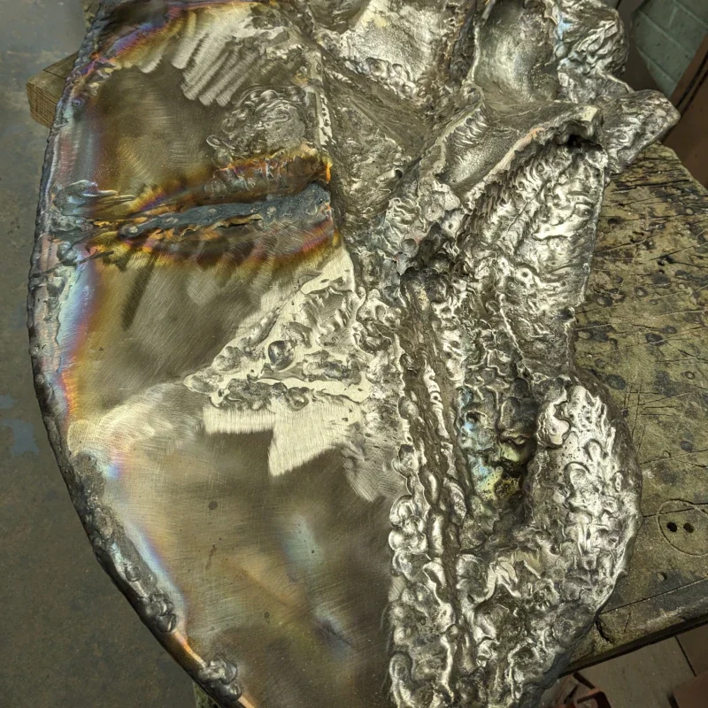 Close-up of a metallic slag or burned metal piece with iridescent and textured surface on a wooden workbench.