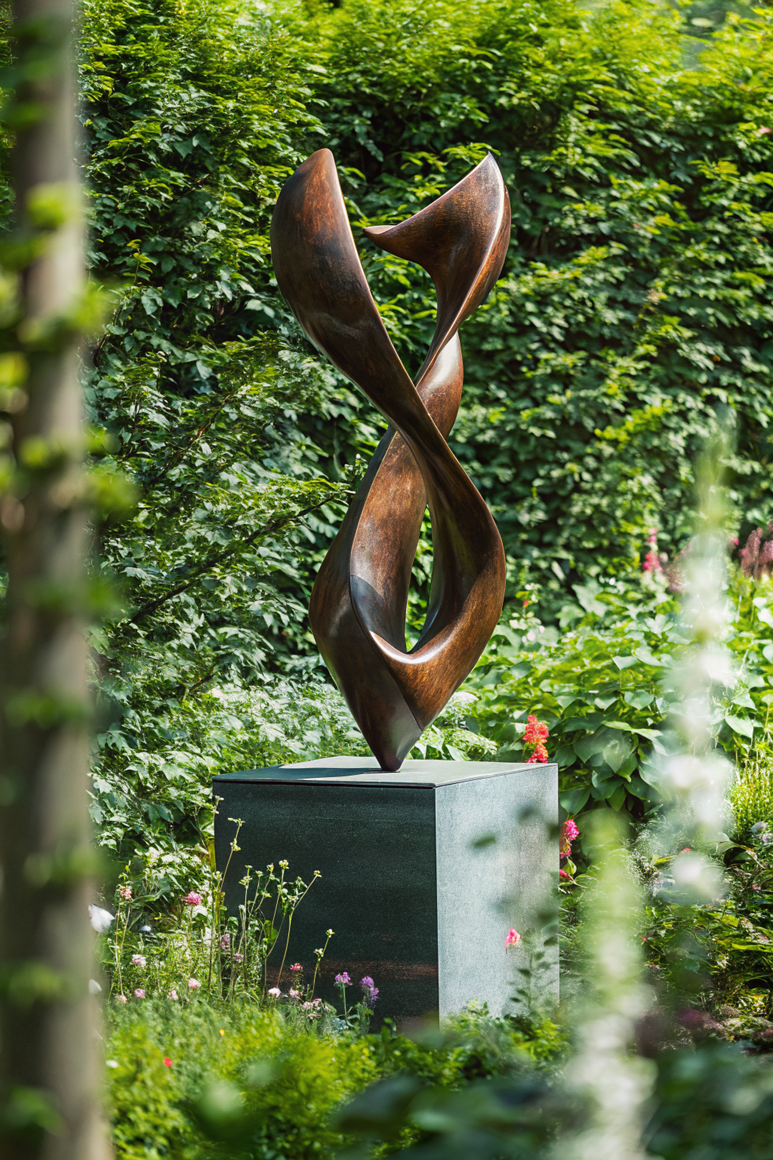 A modern abstract bronze sculpture on a black pedestal is situated in a lush garden with green foliage and colorful flowers.