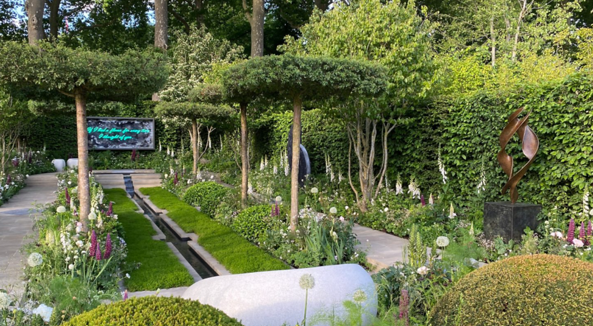 A lush garden with trimmed trees, colorful flowers, and sculptures, including an abstract metal sculpture on a pedestal.