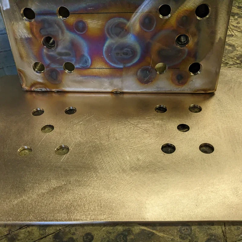 A metal workpiece with multiple drilled holes, placed on a workbench, with a nearby metal sheet that also has drilled holes, possibly indicating metal fabrication or welding preparation.