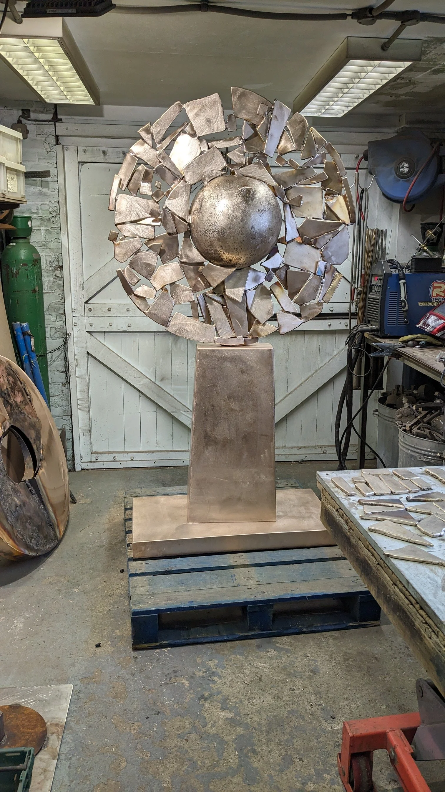 A metal sculpture featuring a large sphere at the center surrounded by broken metal pieces, mounted on a rectangular pedestal in a workshop setting.