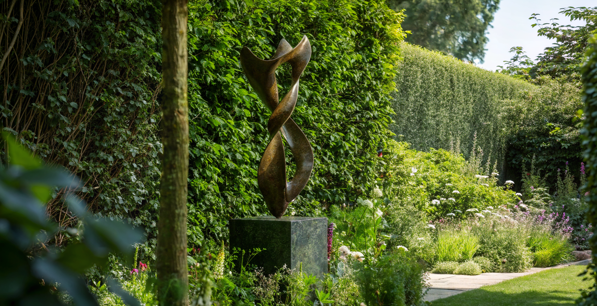 A modern abstract sculpture on a black pedestal in a lush garden with various green plants and trees.