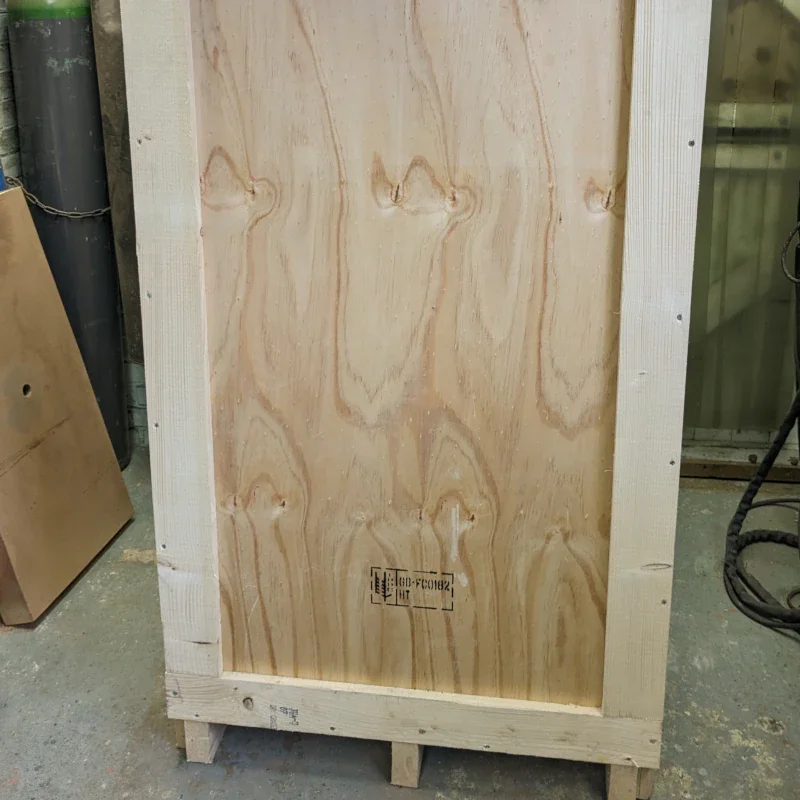A wooden crate with plywood sides and a partly visible package label on the front, in a workshop setting.