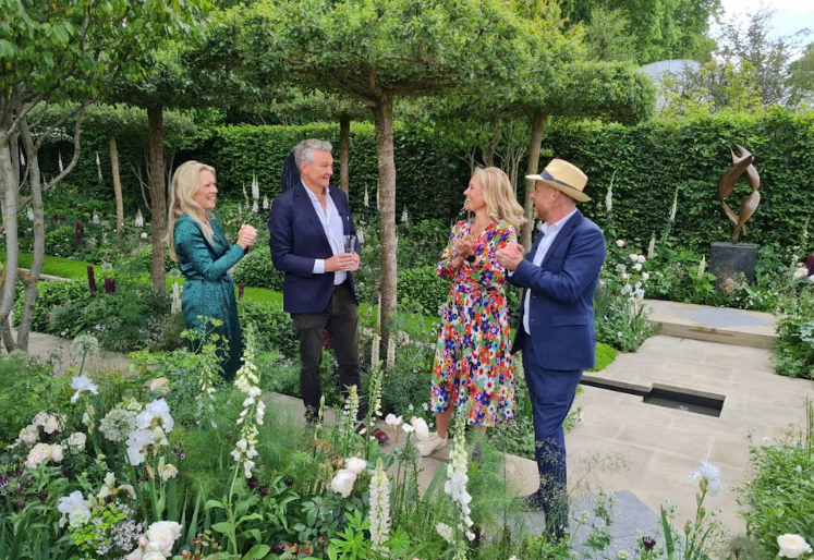Four people, two women and two men, standing and talking in a lush garden with trees, flowers, and sculptures, dressed in formal and colorful attire.
