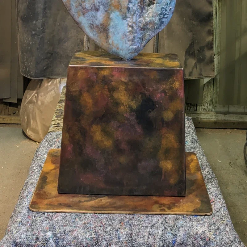 A square black box with colorful abstract patterns sits on top of a larger panel with similar colors, placed on a rough fabric surface.