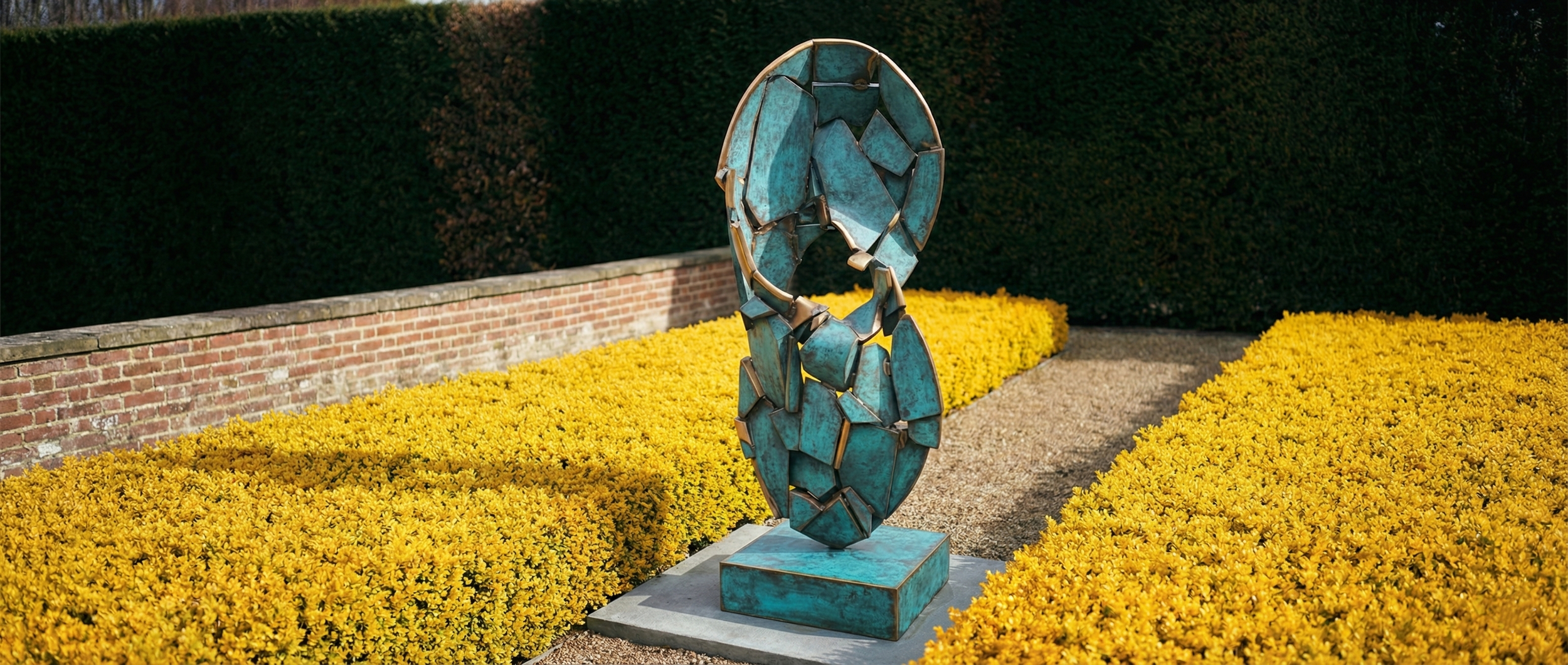 Abstract blue and bronze sculpture on a rectangular base, surrounded by yellow flowers and green hedges.