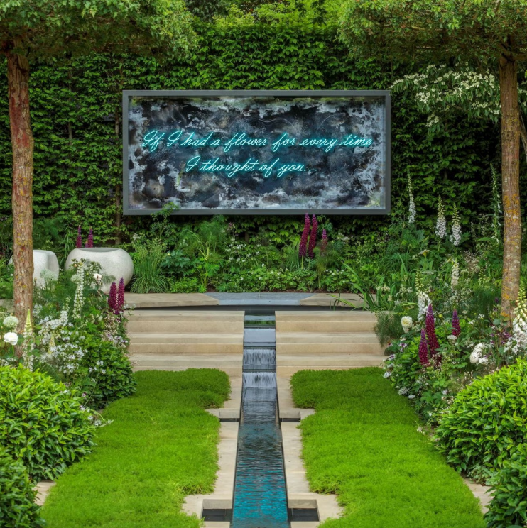 A garden with green grass, lush bushes, and flowering plants, leading to a small waterfall and an illuminated sign with a romantic quote about flowers and thoughts.