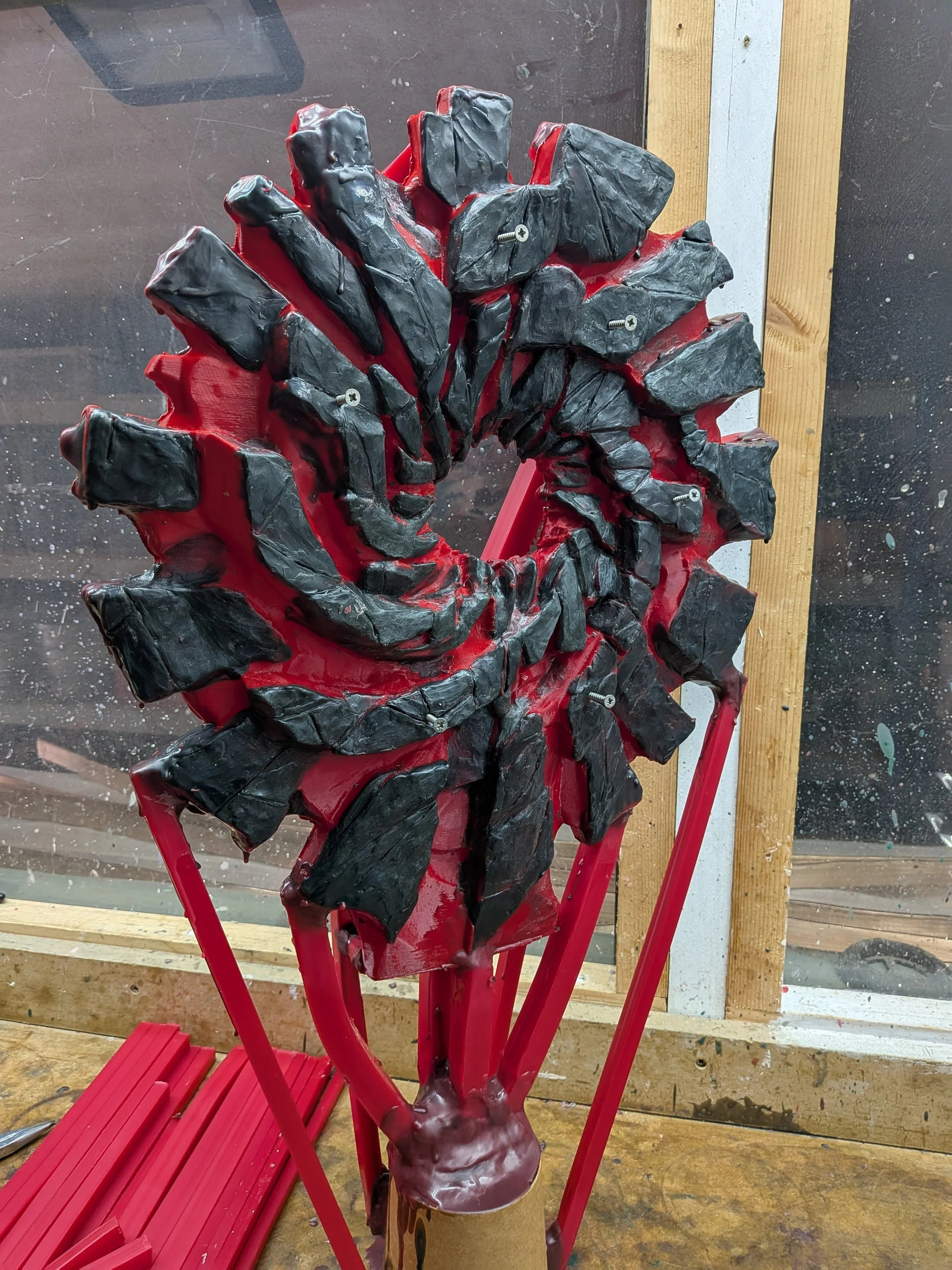 Sculpture of a human face with black rocks and red elements, mounted on a red stand in an artist's workshop.