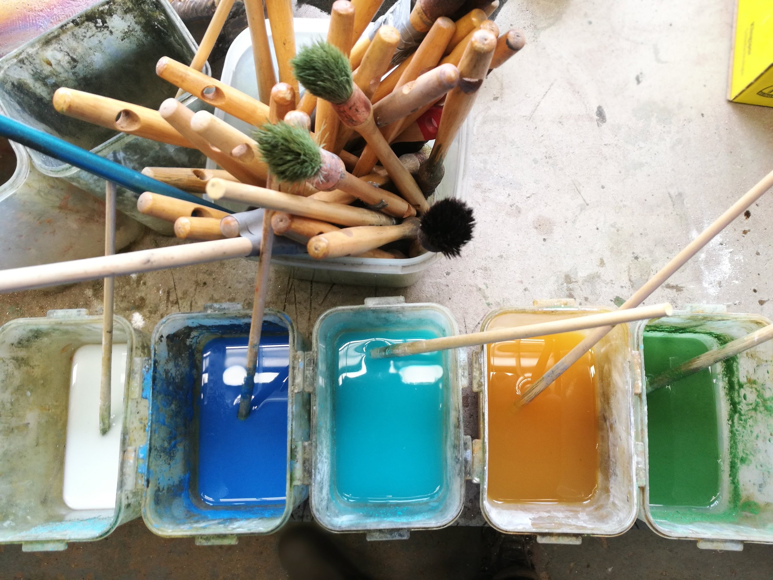 Assorted paintbrushes with wooden handles and bristles, some with green or black tips, resting in trays of various colored paints and liquids on a dirty work surface.