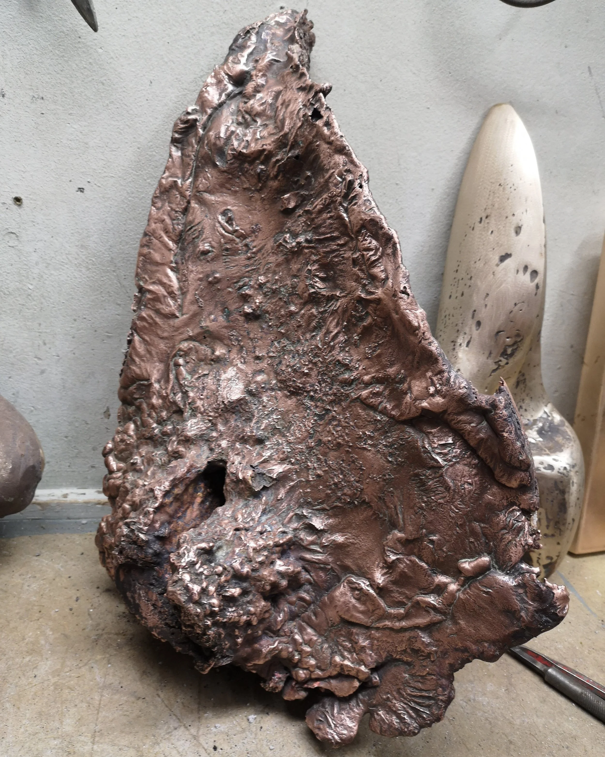 Large abstract sculpture made of a textured, reddish-brown material, resembling rock or metal, placed on a concrete floor.