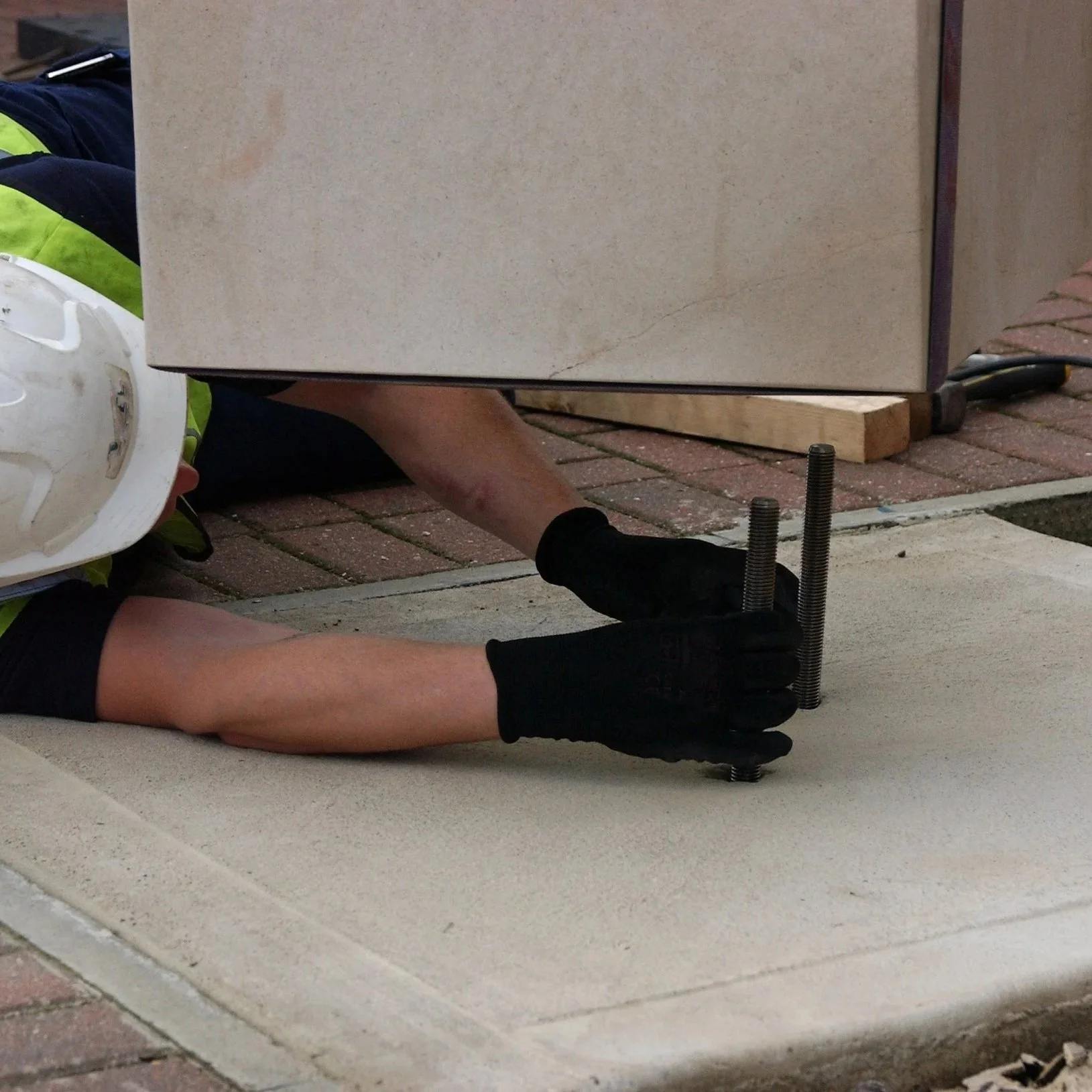 Worker installing a concrete slab with large bolts using a level, wearing a white helmet, gloves, and safety vest.