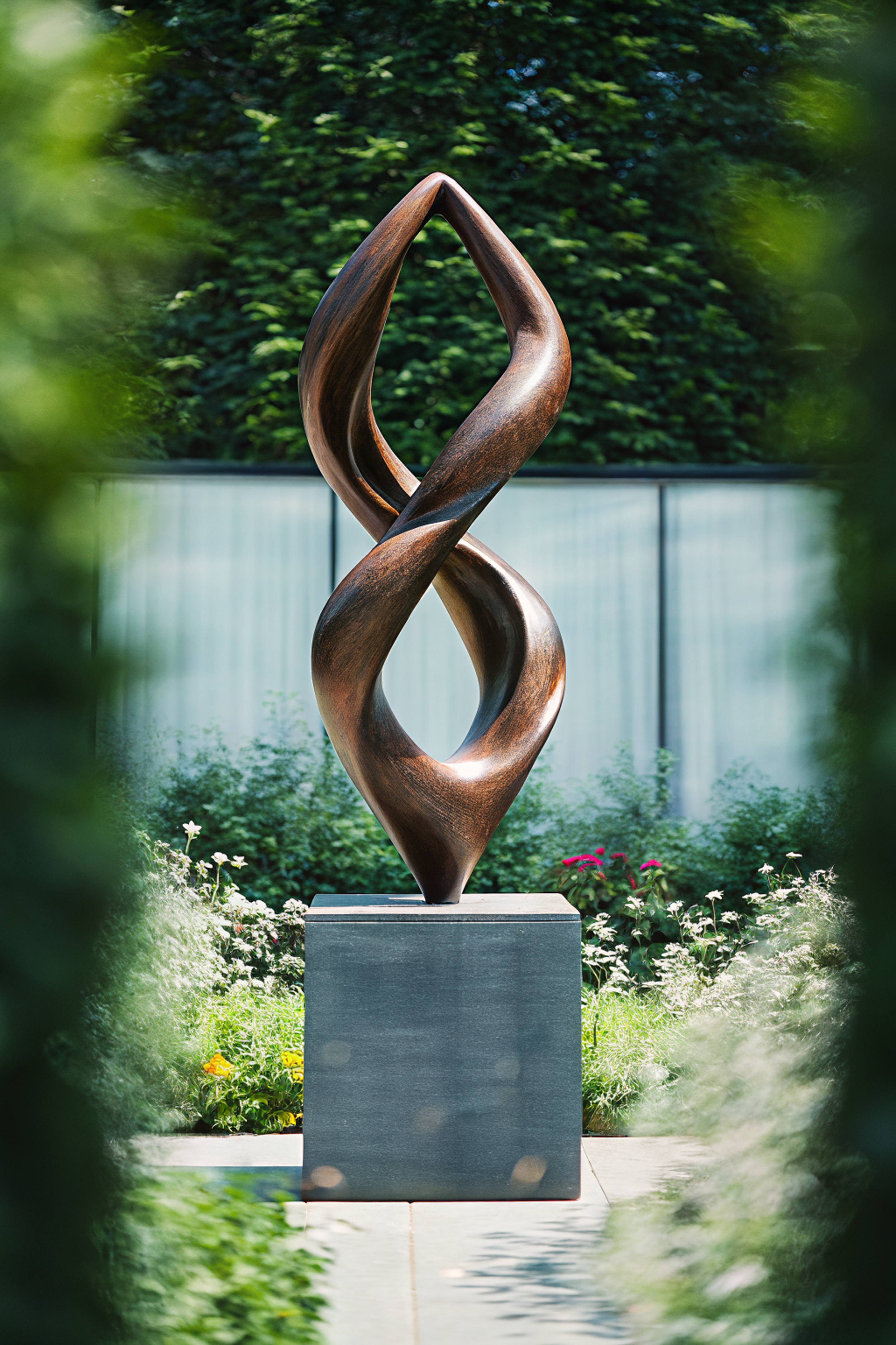 A tall, abstract wooden sculpture with intertwined, twisting shapes on a stone pedestal in a garden with green bushes and trees in the background.
