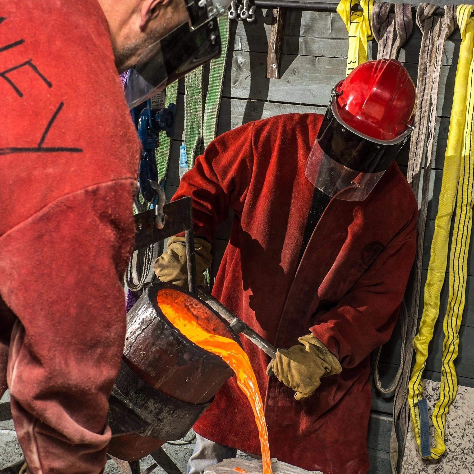 Two blacksmiths working with molten metal; one pouring molten metal from a crucible, both wearing protective gear including gloves, aprons, face shields, and helmets in a workshop setting.