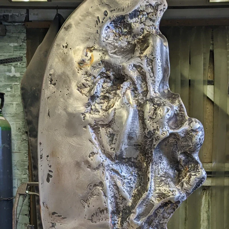 Close-up of a metal sculpture resembling a human face in profile, with detailed textures and indentations.