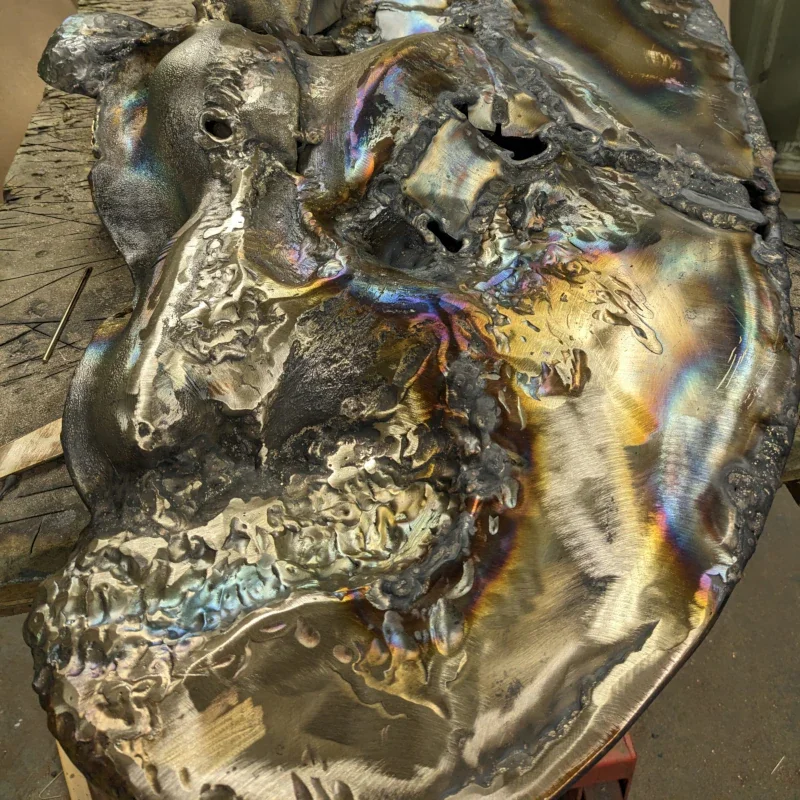 Close-up of a melted, irregularly shaped metal sculpture with rainbow-like burn marks and textured surfaces.
