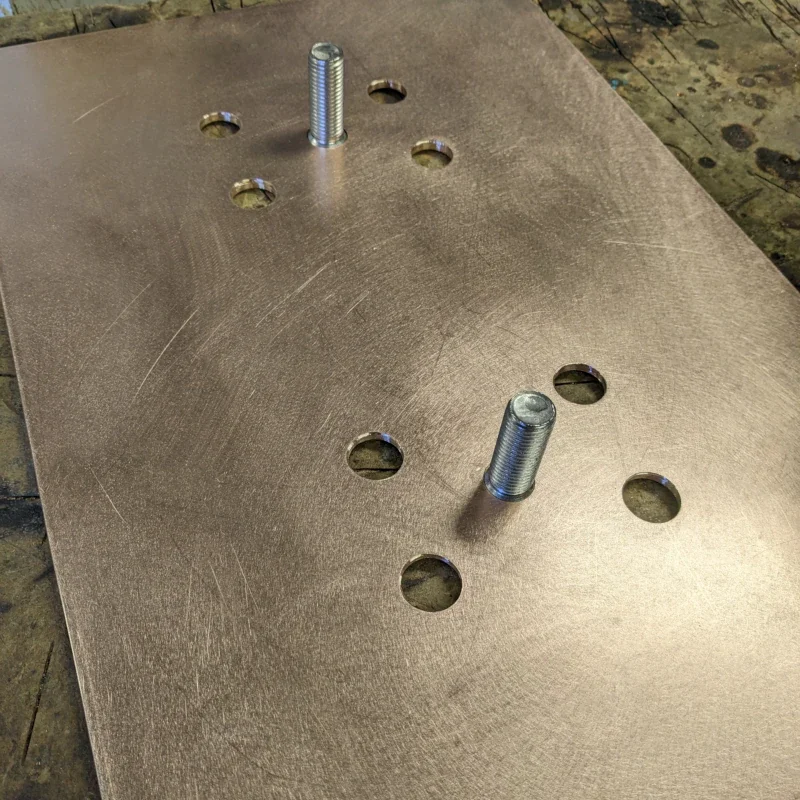 A metal plate with six drilled holes and two protruding threaded bolts, resting on a workbench or table.