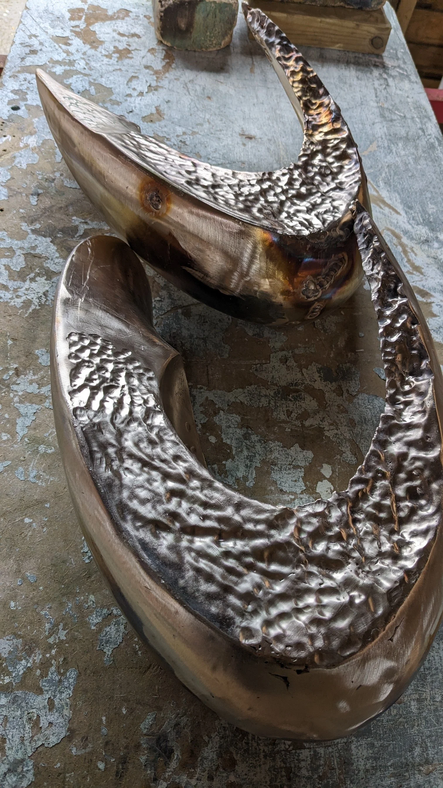 A large metallic sculpture with a curved, crescent shape and textured surface, placed on a weathered wooden surface.