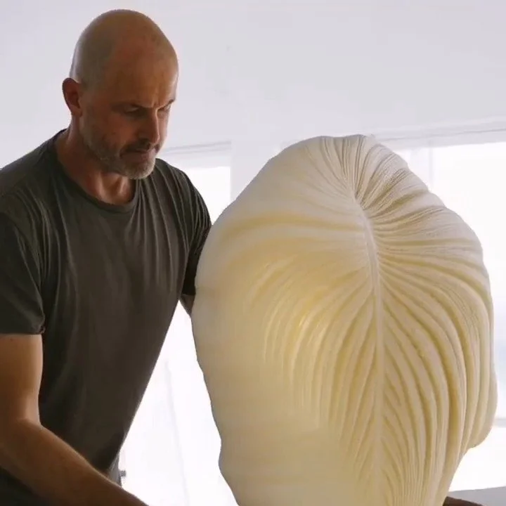 A man examining a large, artfully carved sculpture made of layered, pale material that resembles hair or fabric, in a bright room.
