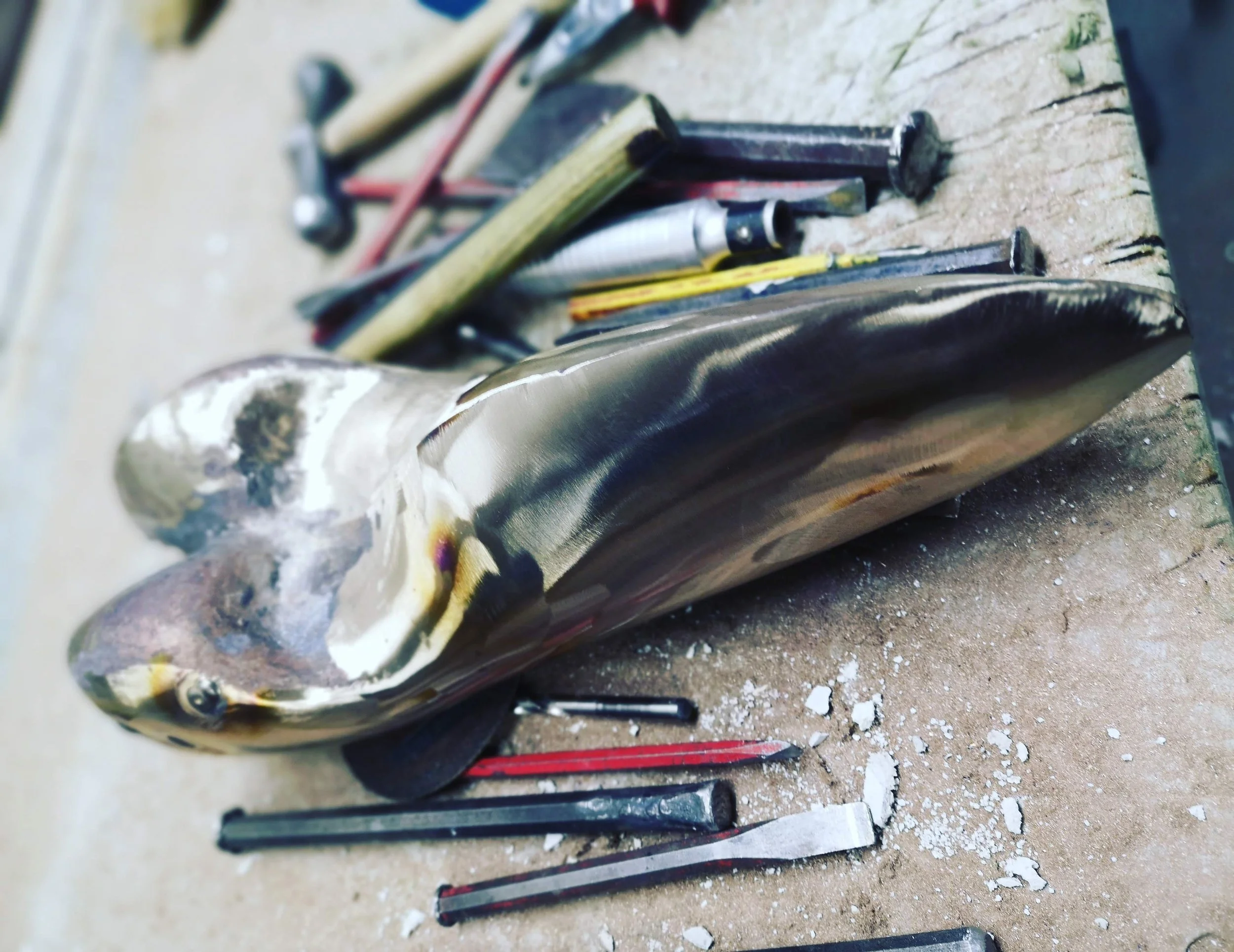 Close-up of a metal sculpture resembling a fish with colorful details, lying on a workbench among tools and debris.