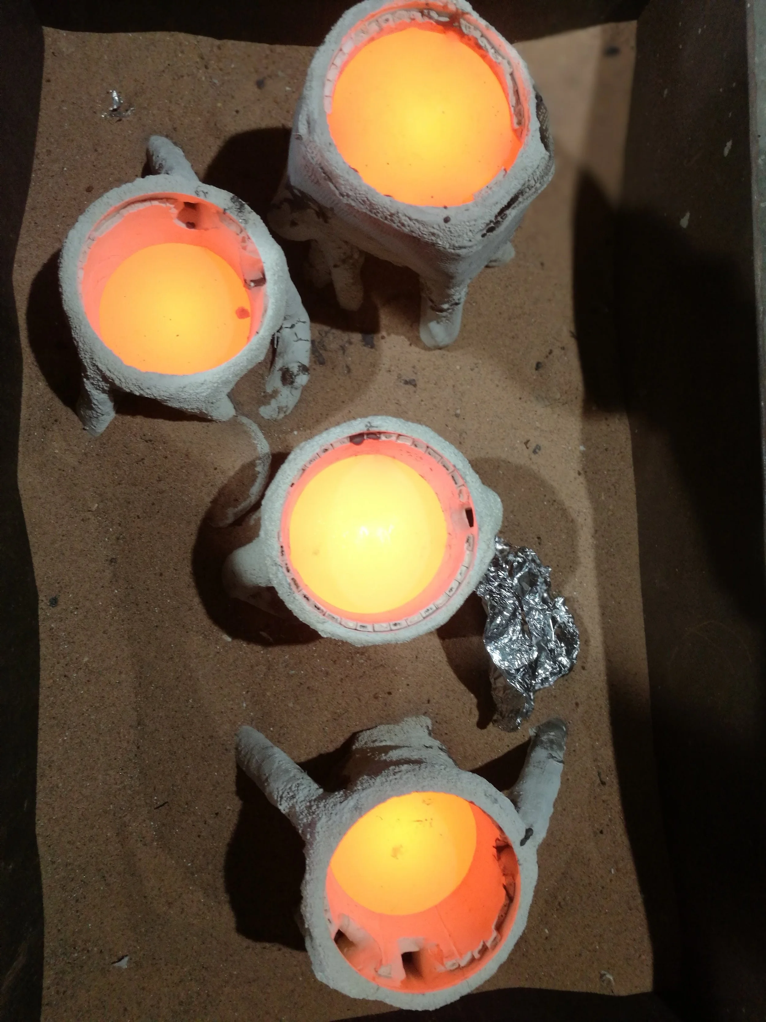 Four hollowed-out eggshells with glowing orange light inside, arranged on a sandy surface, some with remnants of painted or decorated shells and foil underneath.