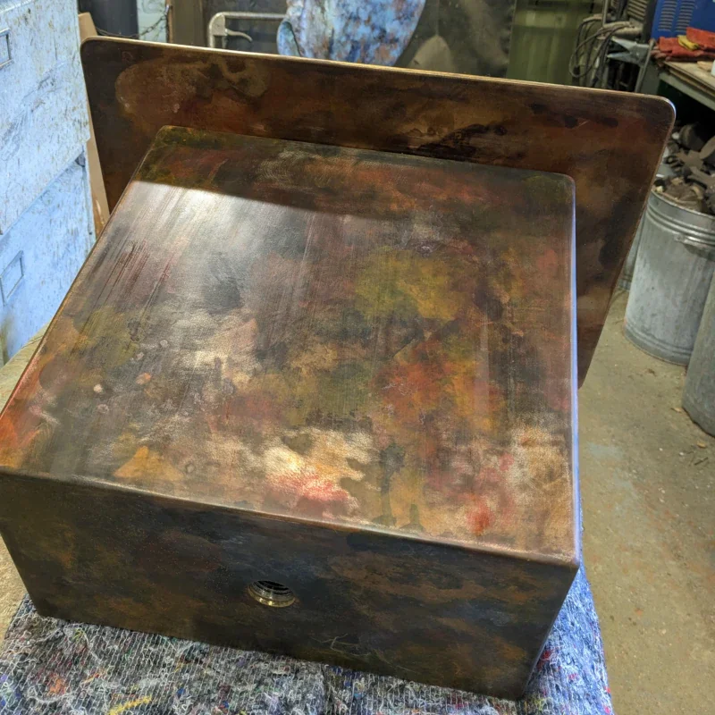 A metal box with a tarnished, multicolored patina surface, resting on a multicolored cloth in a workshop environment.