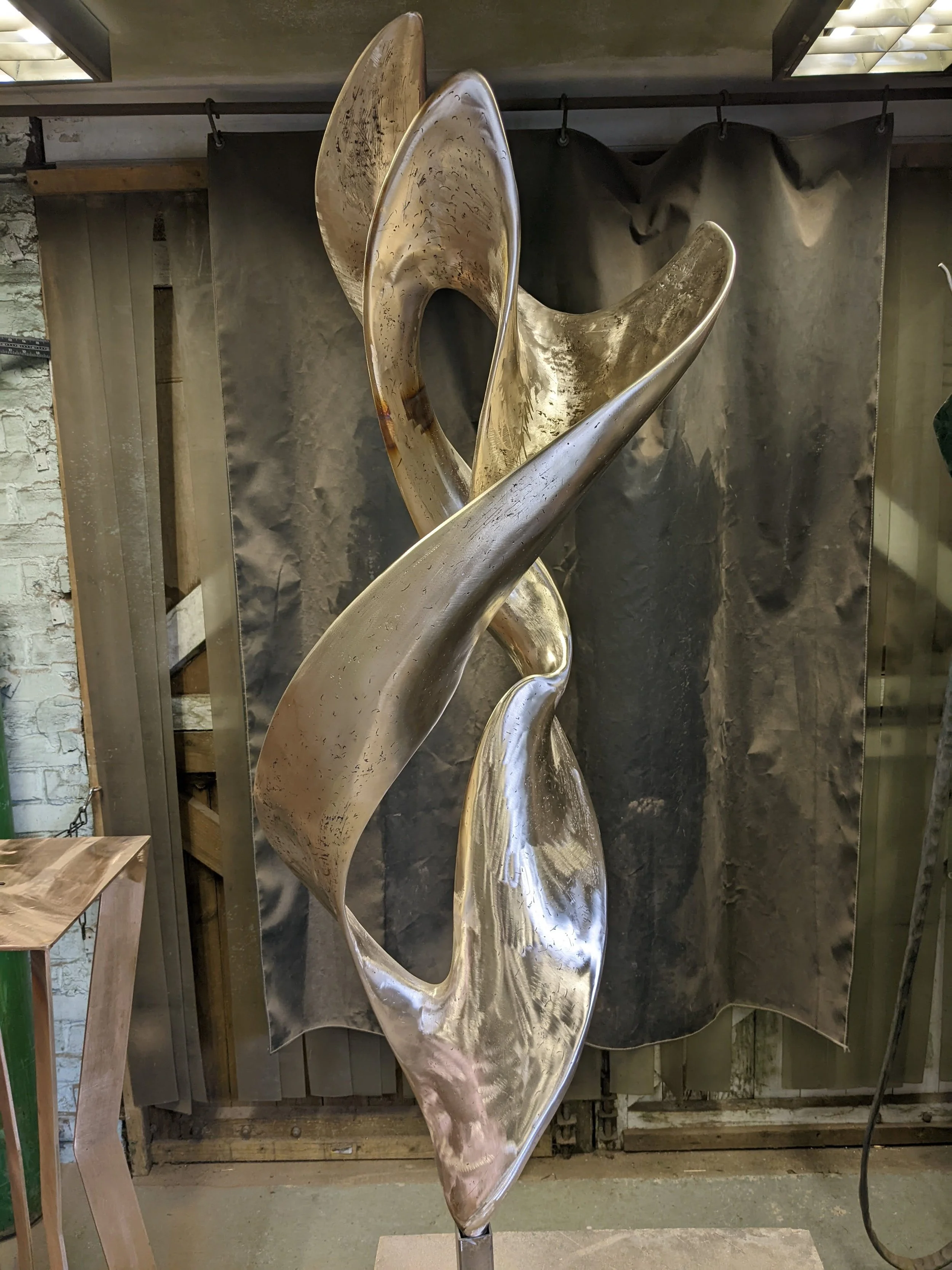 A large, abstract metallic sculpture with flowing, curved shapes. The sculpture is mounted on a small pedestal and is situated in an art studio or gallery space with unfinished walls and a black backdrop.