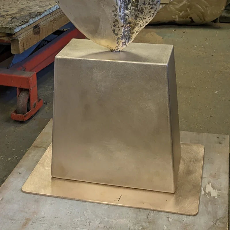 Metalworker shaping a large metal cube with a hammer on top in a workshop.
