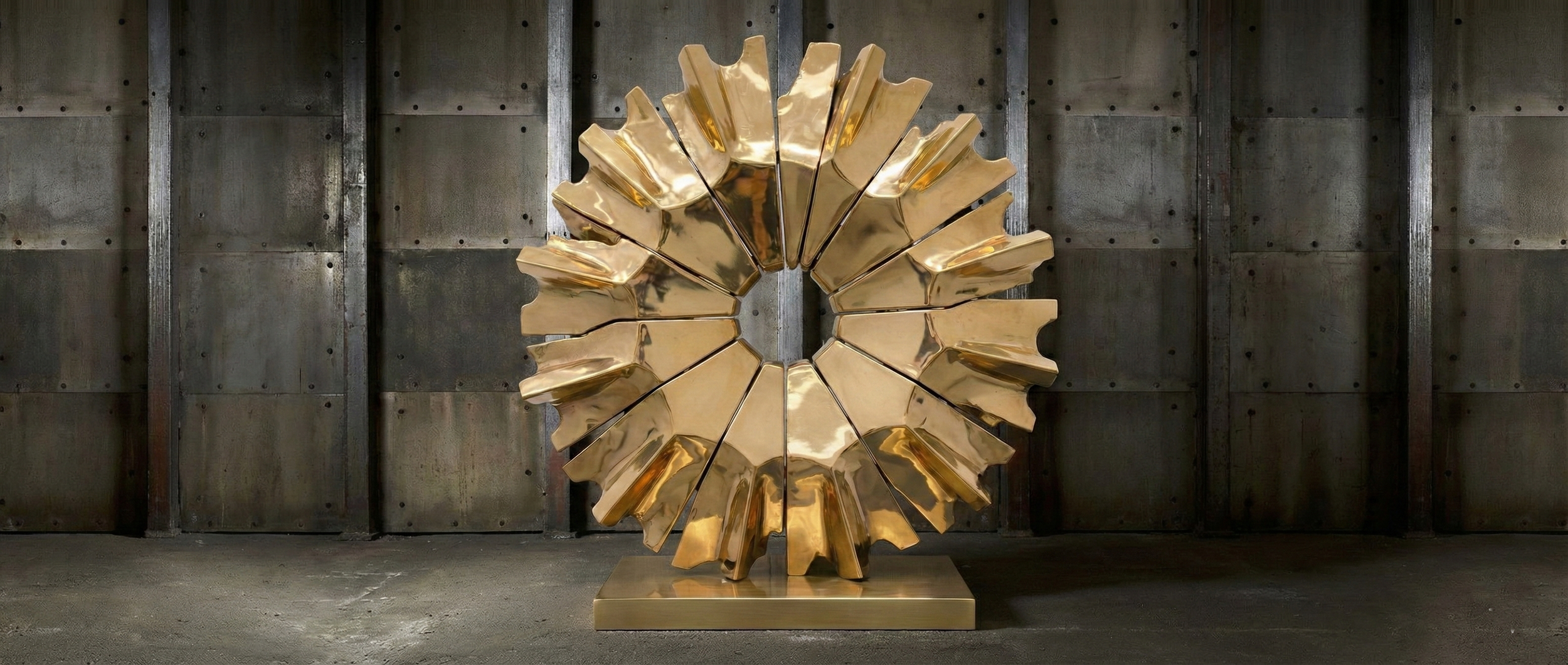 A large, modern gold sculpture resembling a circular gear or sunburst, positioned on a rectangular base against a dark, industrial-style wall with vertical metal beams.