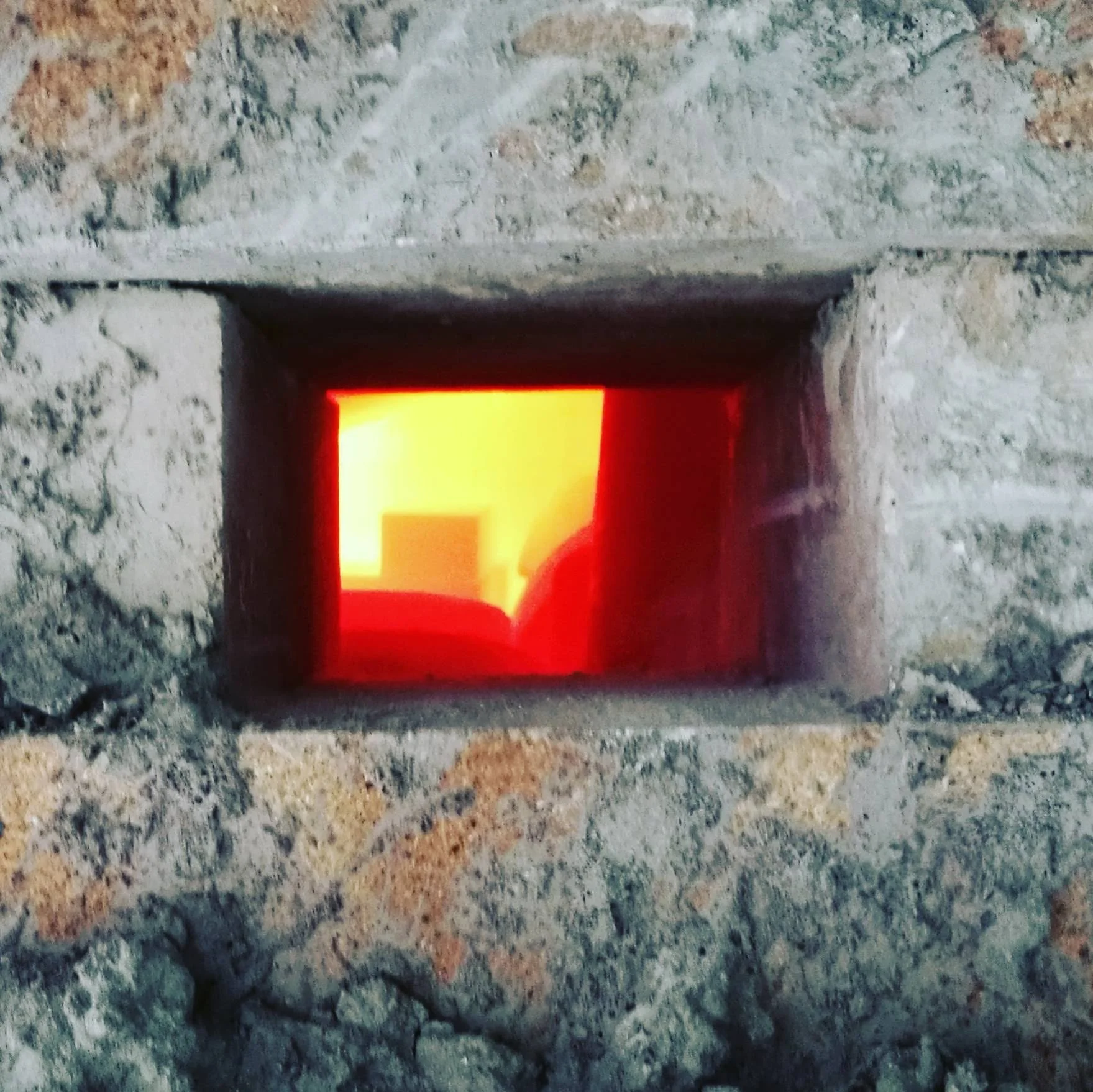A rectangular opening in a stone wall with a bright orange-red light shining through, illuminating the inside.