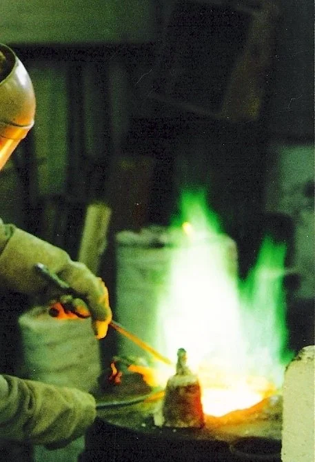 A person working in a metal foundry, pouring molten metal into a mold with green flames visible in the background.